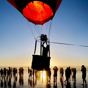 A dramatic image showing a hot air balloon ascending against a vibrant, colorful sky during sunset or sunrise. The balloon's envelope is a striking red color, and its silhouette stands out clearly as it rises up. In the foreground, a group of people can be seen as silhouettes against the warm, golden light, suggesting they are spectators or participants in the hot air balloon experience. The scene evokes a sense of wonder and adventure.