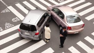 An image showing the aftermath of a car accident at a crosswalk. A white car has collided with a smaller, dark-colored vehicle, and the driver of the dark car appears to be inspecting the damage. There are also people standing nearby, likely responding to the incident. The image conveys a sense of disruption and the potential consequences of a collision in a pedestrian-heavy urban environment.