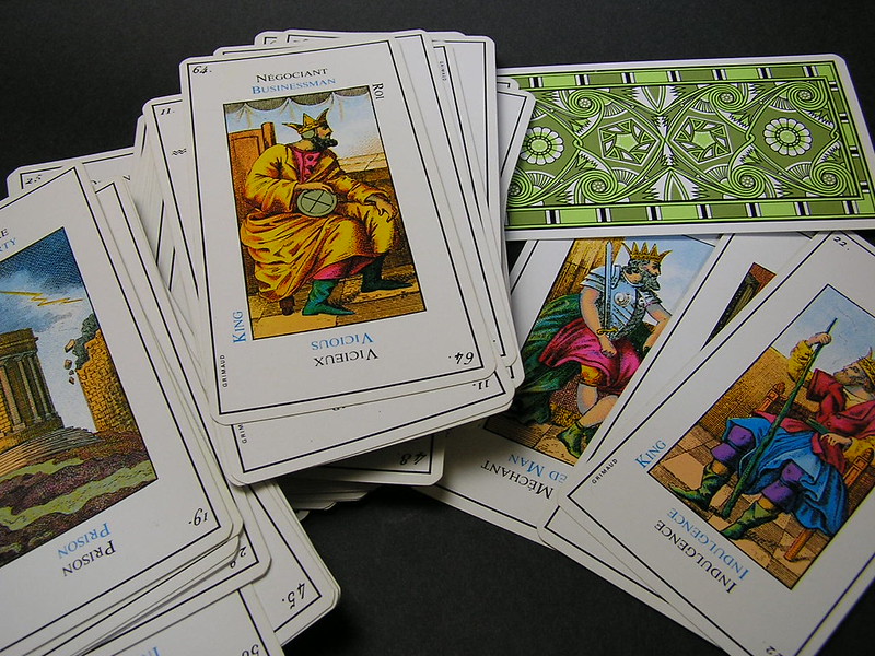 A close-up image of a deck of illustrated tarot or fortune-telling cards spread out on a dark surface. The cards feature vibrant, colorful artwork depicting figures such as kings and scenes with symbolic meanings. The card backs display intricate green and white patterns, adding a decorative element to the set.