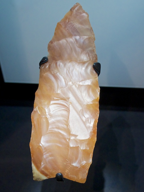 An image of a prehistoric stone tool, likely a flint tool, with a sharp, pointed edge. The tool is crafted through flaking, showing smooth and polished surfaces along its edges. It appears to be a large flake, possibly used for cutting or hunting, displayed in a museum-like setting.