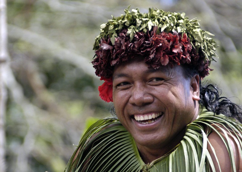 A smiling person wearing a traditional headpiece made of leaves and flowers, paired with clothing or accessories crafted from green foliage. The individual appears joyful, with a vibrant red flower accentuating the headpiece. The blurred background suggests an outdoor, natural setting.