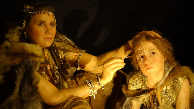 A scene depicting two prehistoric figures, possibly Neanderthals, with one woman applying decorative body paint or adornment to a child’s face. Both figures are wearing fur clothing and jewelry made from beads. The environment suggests a recreation of ancient times, highlighting their cultural practices.