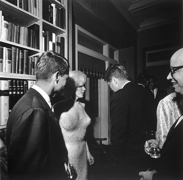 A black-and-white photograph depicts a social gathering in a room lined with bookshelves. The central focus is on a group of elegantly dressed individuals, including a woman in a sparkling dress engaging with two men in suits. The scene has a classic, mid-20th-century ambiance, capturing a moment of conversation and interaction at a sophisticated event.