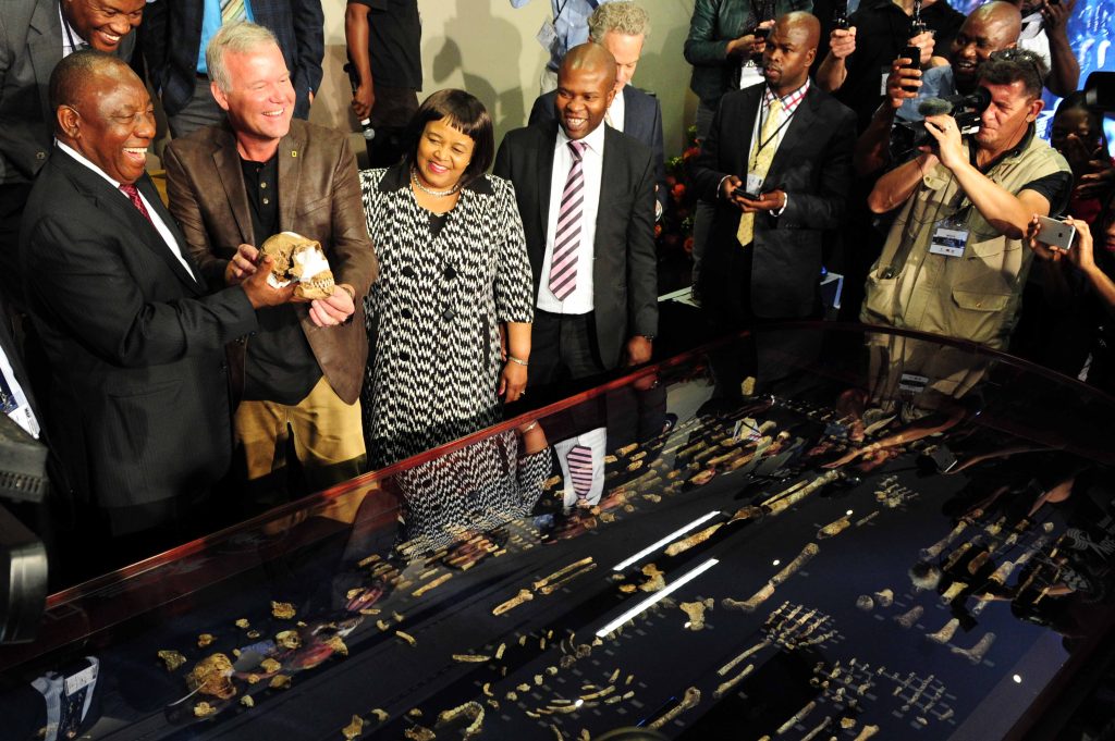A group of people gathered around a display case showcasing ancient artifacts and skeletal remains. In the center, several individuals, including a man holding a skull, stand proudly while smiling. They are surrounded by a group of other people, with cameras capturing the moment. The artifacts displayed in the case include bones and tools, offering a glimpse into an important archaeological discovery. The atmosphere appears celebratory, with the event likely marking a significant historical or scientific achievement.