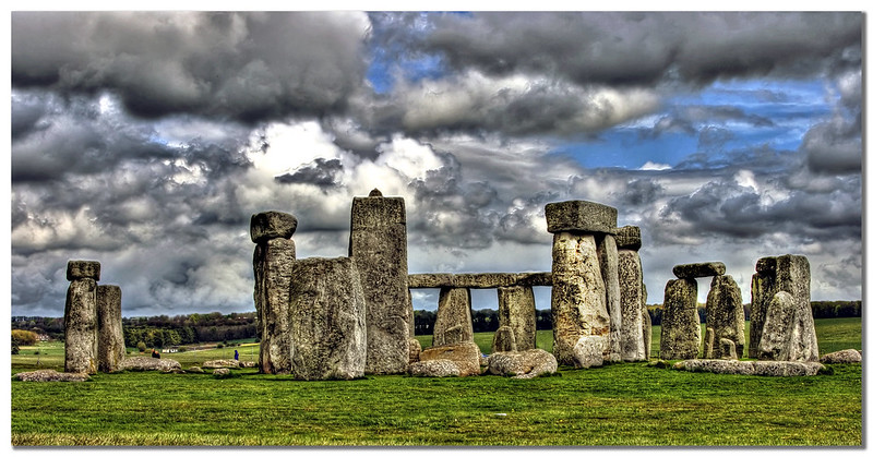A dramatic view of Stonehenge, the ancient megalithic stone circle in England, set against a cloudy and dynamic sky. The large stone structures stand on a vibrant green field, highlighting their imposing and timeless presence. The contrast between the weathered stones and the turbulent clouds adds a sense of mystery and history to the scene.