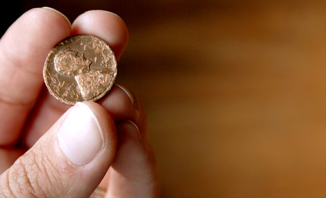 A close-up image of a person's hand holding a heavily worn and damaged coin between their thumb and index finger. The coin appears to be copper-toned, with its surface eroded and markings no longer fully visible. The background is softly blurred, emphasizing the texture and condition of the coin in focus.