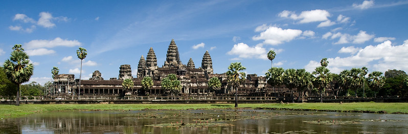 A stunning view of Angkor Wat, the iconic temple complex in Cambodia, surrounded by lush greenery and palm trees. The temple's intricate towers rise majestically under a bright blue sky dotted with white clouds. In the foreground, a reflective water body adds depth and serenity to the scene.
