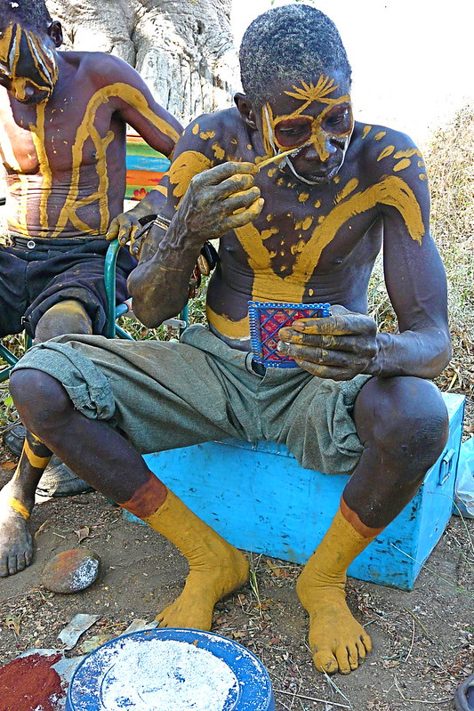 Two individuals are seated outdoors, adorned with intricate body paint in yellow patterns. The man in the foreground applies paint to his face using a thin stick while holding a small hand mirror. He wears rolled-up shorts, and his feet are painted yellow to match the body art. The setting includes a blue box as a seat, a plate with paint materials on the ground, and natural vegetation in the background. The person beside him is also painted, leaning slightly forward.