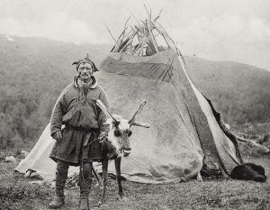 A vintage photograph depicting a Sámi man standing next to a reindeer in front of a traditional Lavvu tent. The man is dressed in traditional Sámi clothing and holds the reins of the reindeer. The surrounding landscape features open tundra with hills in the background, creating a serene, pastoral scene.