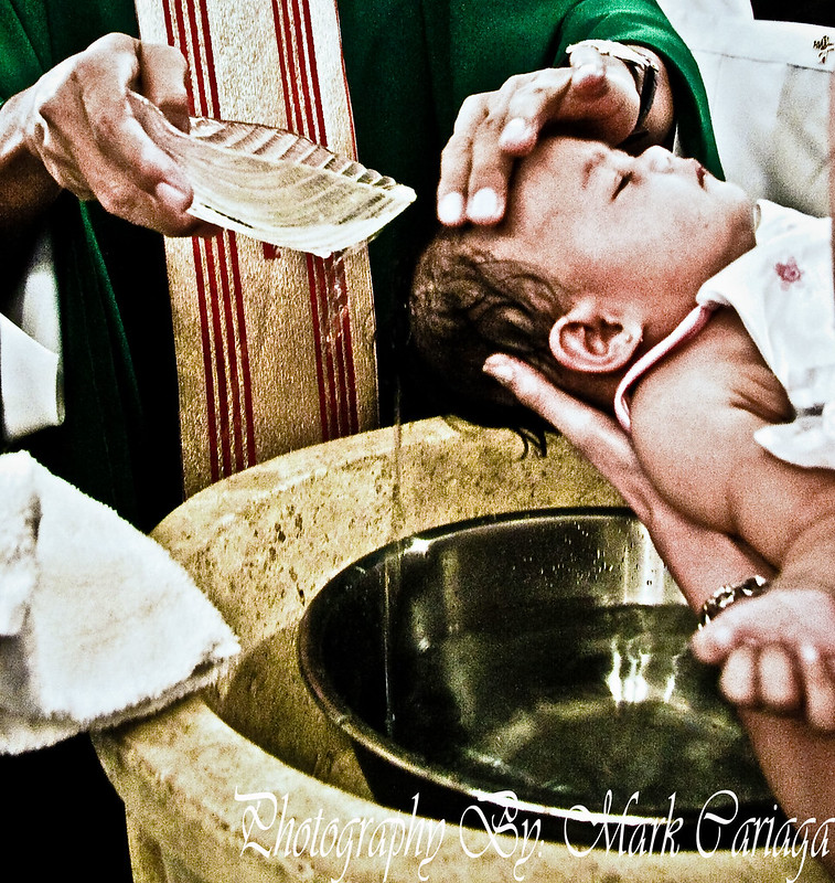 A baby is being baptized in a religious ceremony. A priest in green and white robes pours water from a dish onto the baby's head while gently supporting it with one hand. The scene captures the baptismal font, the baby in white clothing, and the solemnity of the ritual. The atmosphere reflects a sacred and significant moment.