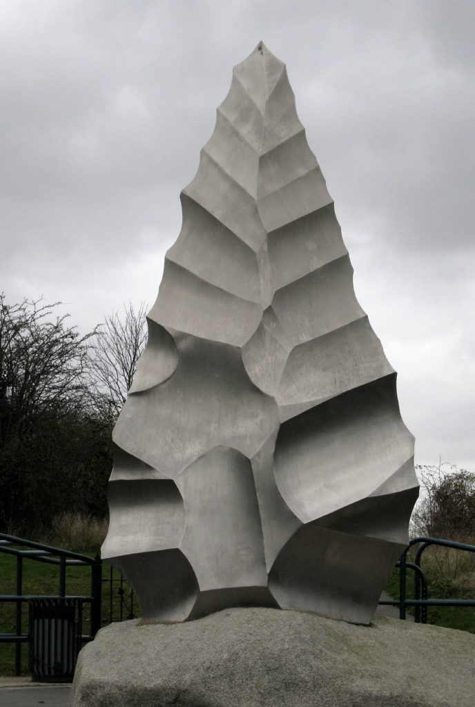 A large metallic sculpture of a flint stone tool resembling an ancient stone blade, positioned outdoors against a cloudy sky. The sculpture has jagged, angled sections that mimic the shape of a prehistoric hand axe, demonstrating the evolution of tools from primitive stone implements to modern artistic representations.