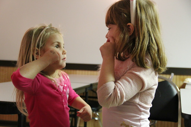 This image shows two young girls engaging in a playful or thoughtful interaction. Both are standing indoors, facing each other, with one gesturing with her hand on her cheek. The girl on the left, dressed in a pink top, looks intently at the other girl, who is wearing a pale pink shirt and has a headband in her hair. The setting appears to be casual, likely a classroom or playroom, with simple chairs and tables in the background. Their expressions convey curiosity and engagement, capturing a moment of connection and communication.