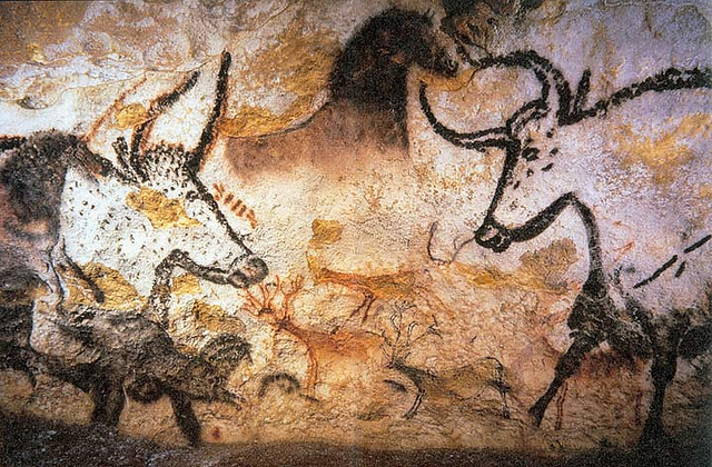 A photo of prehistoric cave paintings depicting animals, likely from the Paleolithic period. The images show bison, horses, and other wildlife drawn with black and ochre pigments on the cave wall, showcasing early human artistic expression. The scene captures motion, with some animals appearing to move or interact with one another.