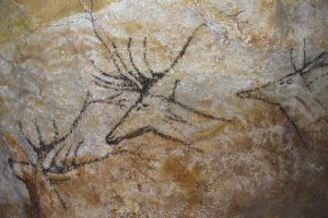 An image of prehistoric cave paintings depicting animals, likely from the Paleolithic period. The artwork shows deer with detailed antlers, drawn with black pigments on the cave wall, highlighting early human efforts in representing the natural world.