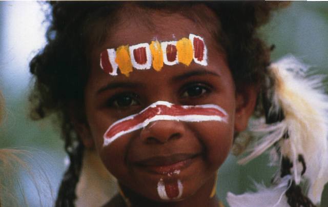 This image captures a young child with a joyful expression, adorned with traditional face paint in vibrant colors of white, red, and yellow. The symmetrical patterns across their forehead, cheeks, and nose appear to hold cultural or ceremonial significance. Their hair is styled naturally, with decorative feathers framing their face. The soft, blurred background hints at an outdoor setting, likely emphasizing their connection to nature and tradition. The image conveys a sense of heritage, pride, and innocence.
