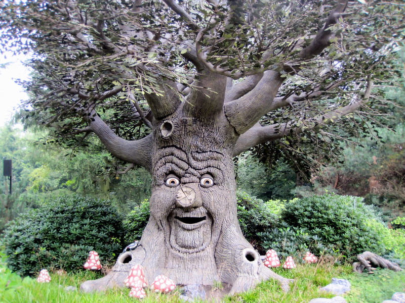A whimsical tree sculpture with a smiling, animated face carved into its wide trunk stands in a lush, green garden. Its branches extend outward, adorned with detailed leaves, giving it a lifelike appearance. Surrounding the tree are clusters of red-and-white polka-dotted mushrooms, enhancing the magical, storybook-like atmosphere. Dense shrubbery and tall trees in the background complete the enchanting scene.