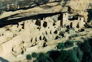 A view of the ancient cliff dwellings at Mesa Verde, showcasing a well-preserved village built into a sandstone alcove. The site features multiple stone structures, including towers and kivas, surrounded by rugged terrain. Visitors can be seen exploring the area, giving scale to the remarkable architectural achievement of the Ancestral Puebloans.
