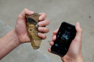 A person holds two objects side by side, a stone tool and a modern smartphone. One hand grips a prehistoric flint stone, and the other holds an iPhone displaying the time. The contrast between the ancient tool and the modern technology symbolizes the evolution of human tools and technology over time.