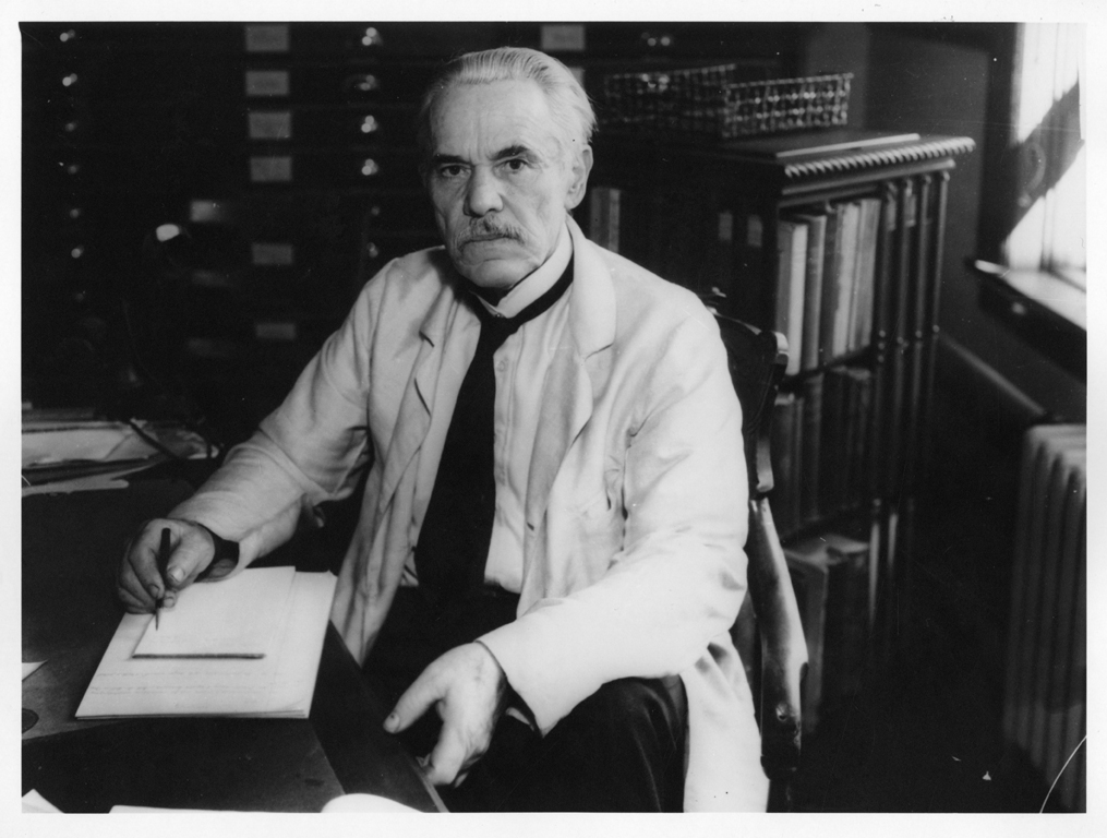 A black and white photograph of a man sitting at a desk, wearing a white lab coat and a tie. He is holding a pen and writing on a piece of paper, looking directly at the camera with a serious expression. Behind him, there are shelves with books and filing cabinets. The image appears to be from an earlier era, possibly the mid-20th century.