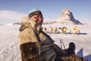 A person dressed in warm fur clothing smiles while riding a dog sled across a frozen landscape. In the background, a team of sled dogs stands on the snow with a rugged, snow-covered mountain rising prominently under a clear blue sky.