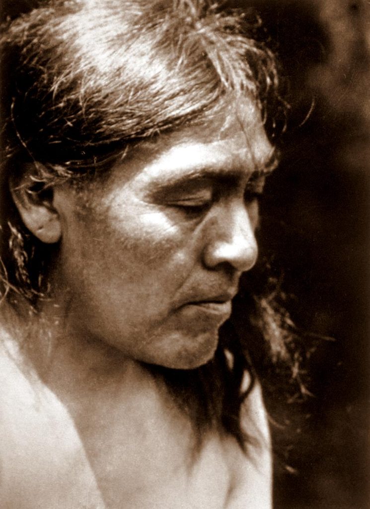 A sepia-toned portrait of a Native American individual with long hair, looking downward in a contemplative pose. The lighting highlights the facial features, creating a sense of depth and emotion. The background is softly blurred, keeping the focus on the subject.