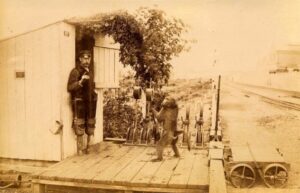 A vintage sepia-toned photograph showing a railway signalman standing at the door of a small wooden signal hut. Next to him, a trained baboon stands on a wooden platform, seemingly assisting with railway operations. The background includes tracks, vegetation, and a distant view of the railway line.