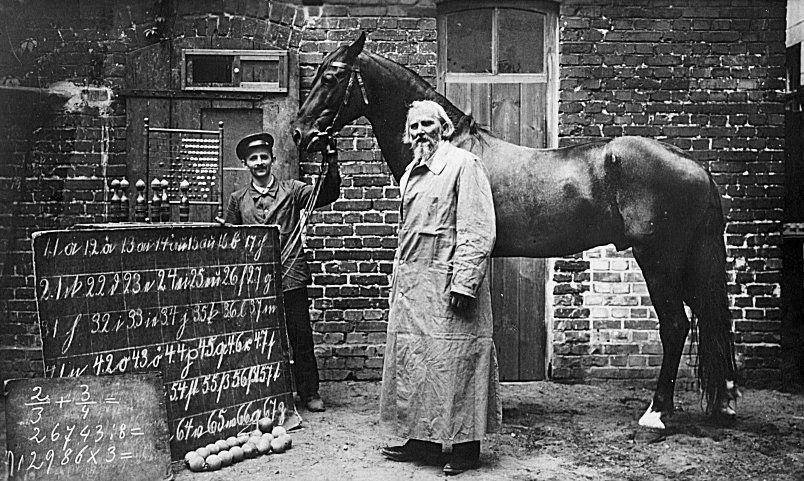 The image depicts a historical scene featuring a man in a long coat standing next to a horse, with another man in uniform holding the horse's reins. They are in a brick courtyard, and a large chalkboard with mathematical equations and notations is prominently displayed in the foreground. Several objects, possibly for demonstrations or experiments, are placed near the chalkboard, adding to the educational or experimental atmosphere.