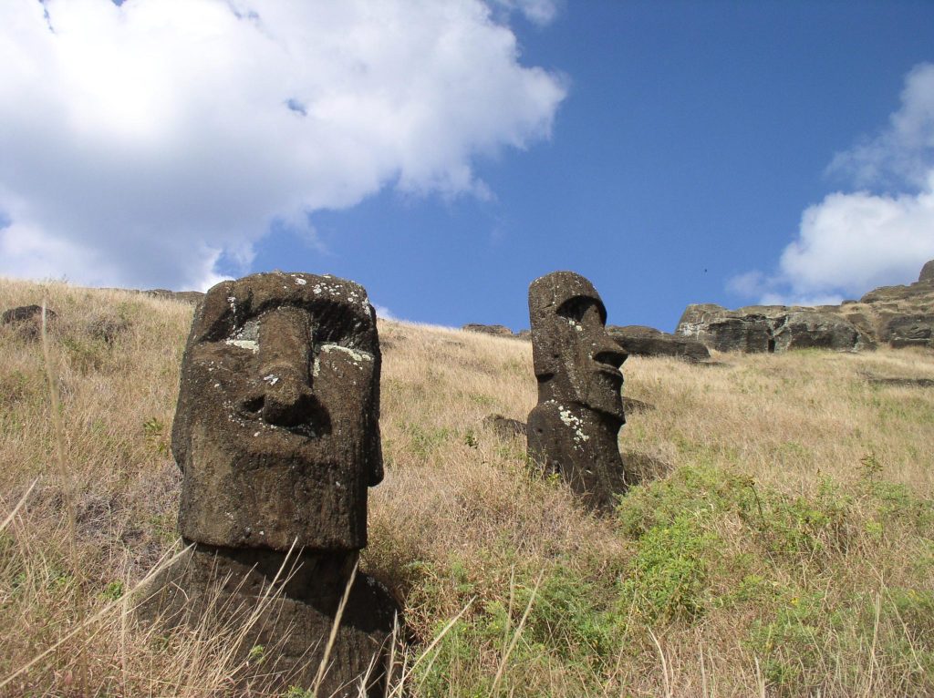 Two iconic moai statues stand on a grassy hillside under a bright blue sky with scattered clouds. The statues, carved from dark volcanic rock, feature elongated faces with prominent noses and deep-set eyes. The surrounding terrain is dry with patches of vegetation, typical of Easter Island's landscape.