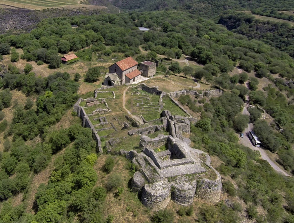 Aerial view of a historic archaeological site, showing the remains of stone structures surrounded by dense greenery. The site includes ancient ruins with well-preserved walls and foundations, and a small building with a red-tiled roof is visible in the center. The surrounding landscape is lush with trees, and there is a road and a bus parked nearby. The area appears to be in a remote, hilly region.