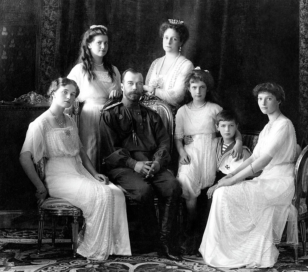 A black-and-white photograph of the Russian imperial family, the Romanovs, featuring Tsar Nicholas II seated in the center with his wife, Tsarina Alexandra, standing beside him. Their children are arranged around them, with the four daughters and son, Alexei, pictured. The daughters are elegantly dressed in white gowns, while Alexei is wearing a sailor suit. The background features rich drapery, and the family poses formally for the photo.