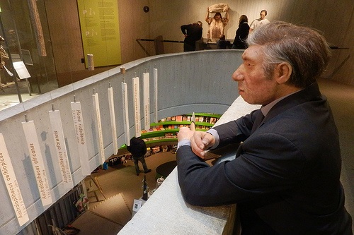 A museum display features a life-sized model of a Neanderthal man dressed in a suit, leaning on a railing and gazing over an exhibit area. In the background, informational panels and a lower level with displays are visible.