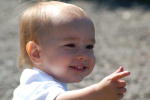 A close-up of a smiling toddler with light hair and wearing a white shirt. The child is pointing forward with their hand, appearing happy and curious. The background is blurred, with a mix of gray and natural outdoor colors.