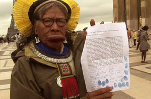 An Indigenous man, wearing a traditional yellow feather headdress, glasses, and cultural jewelry, holds a document with text and blue fingerprints, symbolizing signatures. He stands in a public urban setting with people and buildings in the background, suggesting participation in a formal or advocacy event.