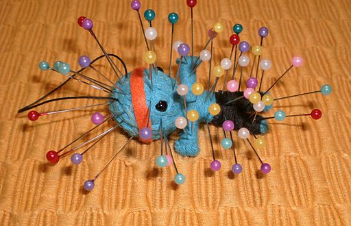 A small blue doll with an orange headband is pinned with multicolored sewing pins, creating a spiky appearance. The doll is placed on a textured yellow-orange surface, and its black details contrast with the vibrant pins scattered across its body.