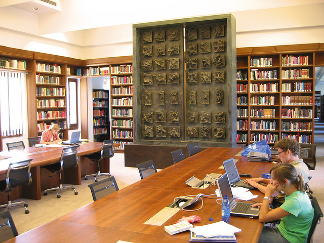 Two students are sitting at a table in a library. They are reading on their laptop computers.