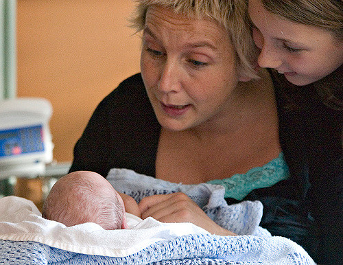 An adult and a child look lovingly at a newborn wrapped in a blue blanket, engaging with the baby through facial expressions and speech.