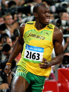 Usain Bolt, wearing a yellow and green Jamaica uniform, sprinting during an Olympic event with photographers in the background.