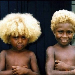 Two young Melanesian children with dark skin and naturally blonde, curly hair, standing against a dark background.