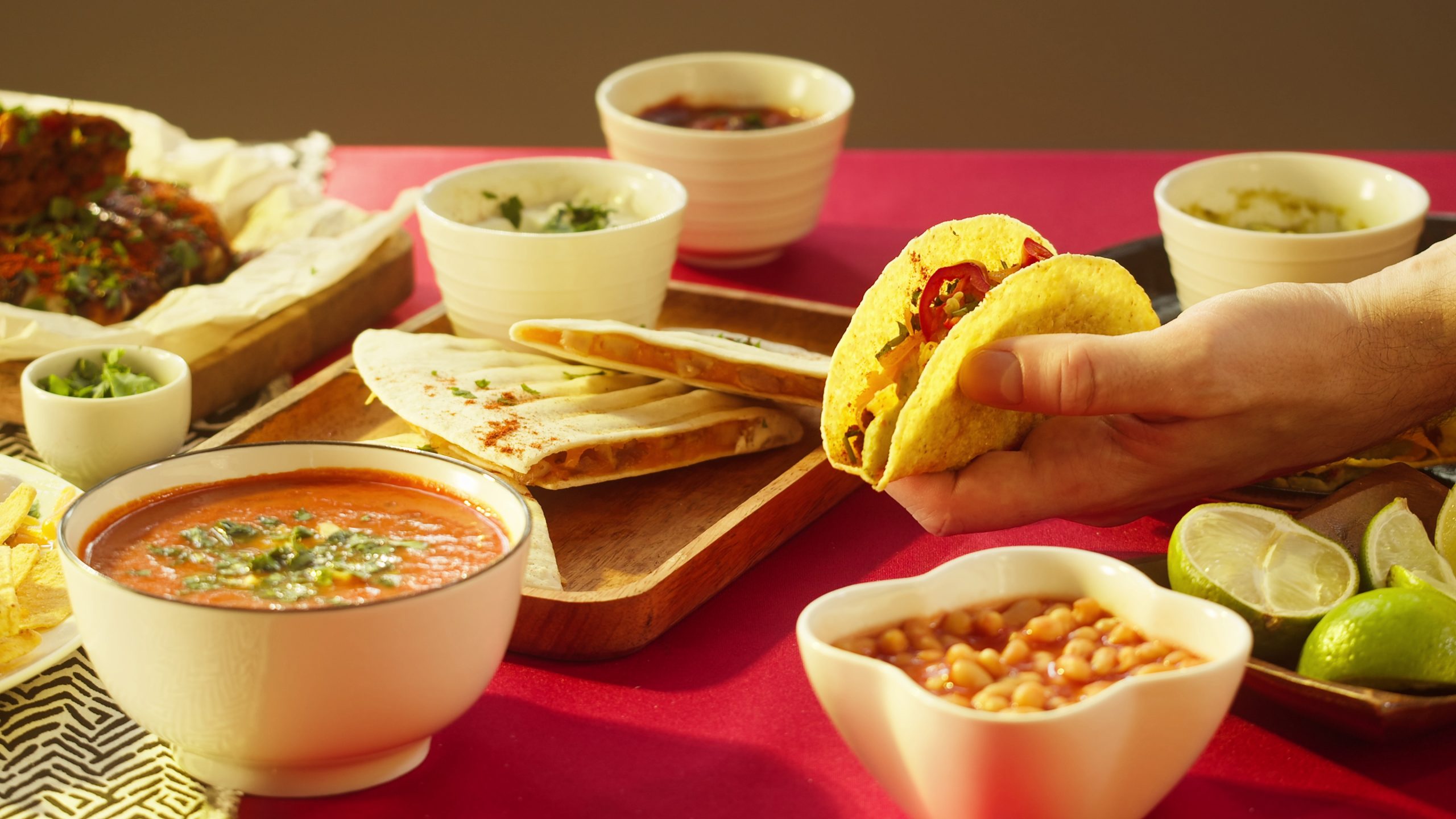 A hand holding a taco filled with vegetables beside a table spread of Mexican food, including quesadillas, salsa, beans, limes, and other small bowls of toppings.