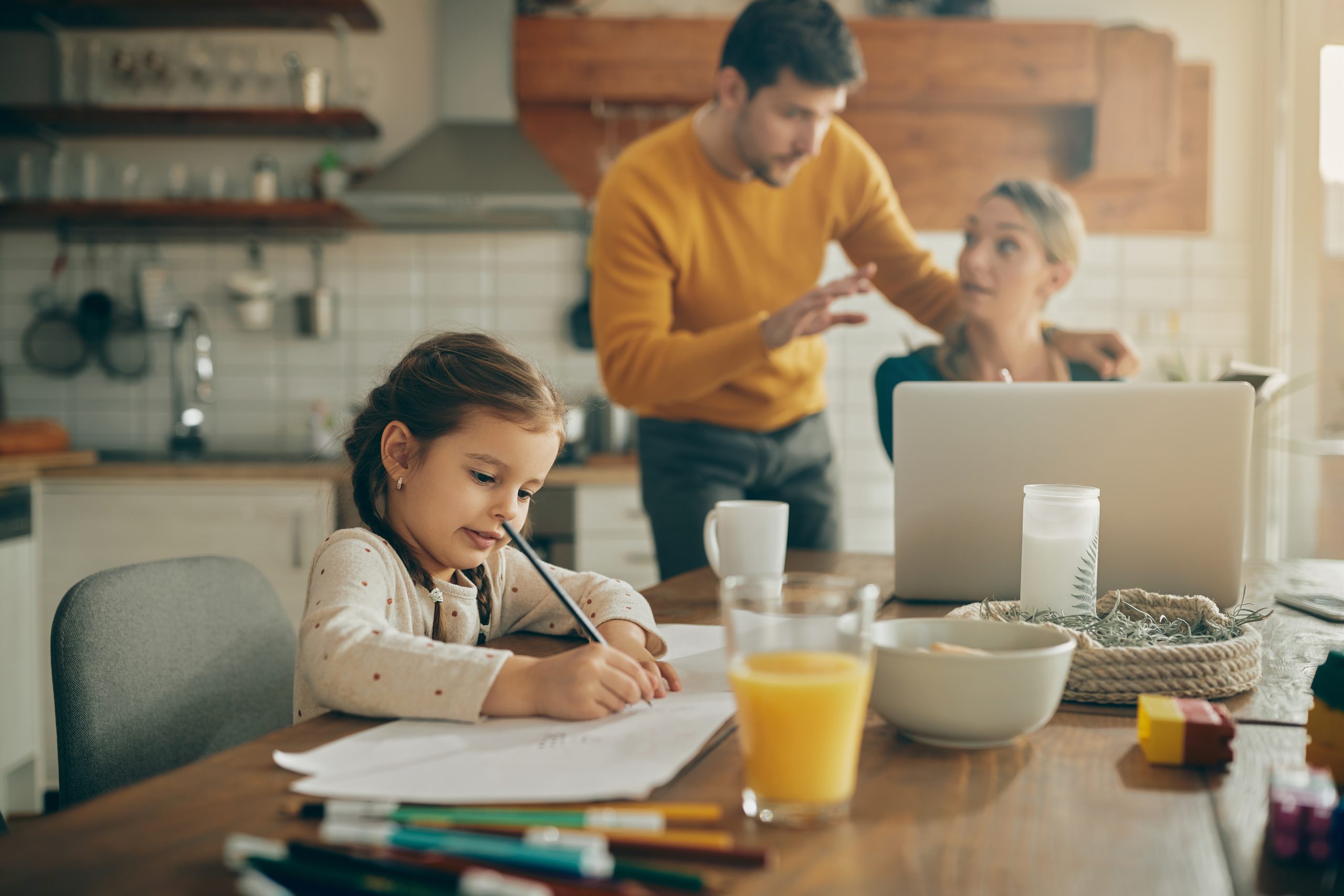 A young girl writes homework at a table while her parents work on a laptop in the background.