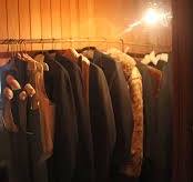 The image shows a dimly lit wooden closet with a series of clothes hanging on a rod. The garments include jackets, coats, and other items on hangers. A single exposed light bulb illuminates the scene, casting a warm glow on the wooden walls and the clothes.