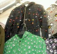 A display of colorful hoodies hanging on a rack. The front hoodie is green with a pattern of white and green mushrooms. Behind it, there is a black hoodie with a pattern inspired by the Pac-Man video game, featuring Pac-Man and ghosts. Additional hoodies with various patterns, including skulls and Hello Kitty, are visible in the background.