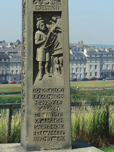 Tall stone monument featuring a dark rectangular plaque with engraved text, positioned on a paved base near a waterfront. Lush grasses and a calm waterway fill the background under a clear sky.