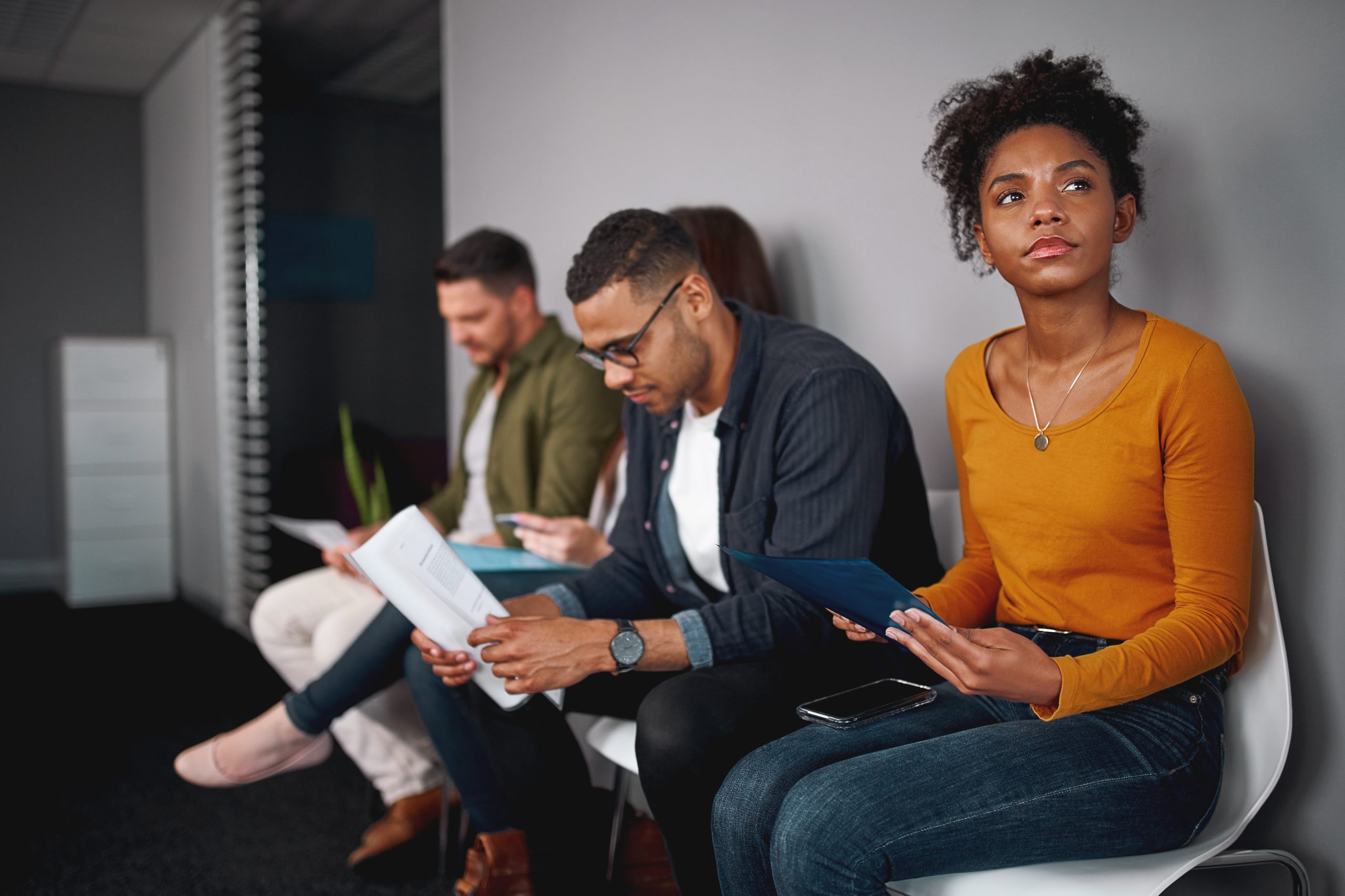 Three individuals sitting in chairs against a wall appearing to line up for an interview. Two men are looking at documents in their hands and a woman is looking up with an inquisitive look on her face. Adobe Stock