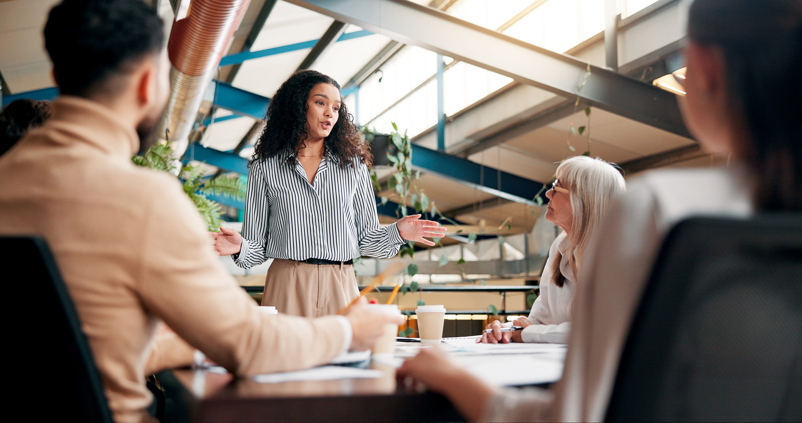 A woman speaker stands in front of a three other individuals who are taking notes at a table. Adobe Stock