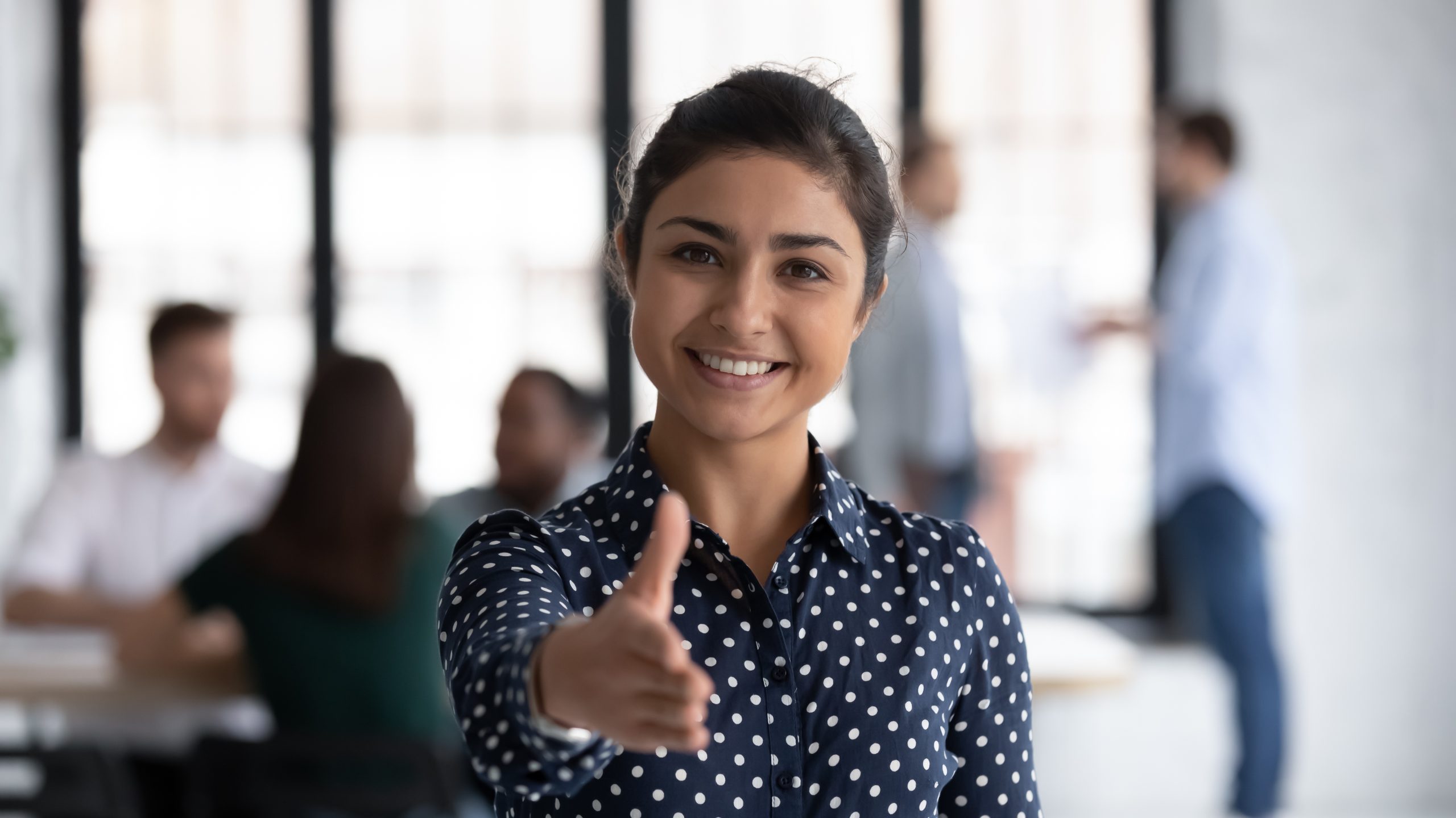 A woman with dark hair with dressed in a blue polka dot professional shirt extending her hand for a handshake with non identifiable people in the background. Adobe Stock Image