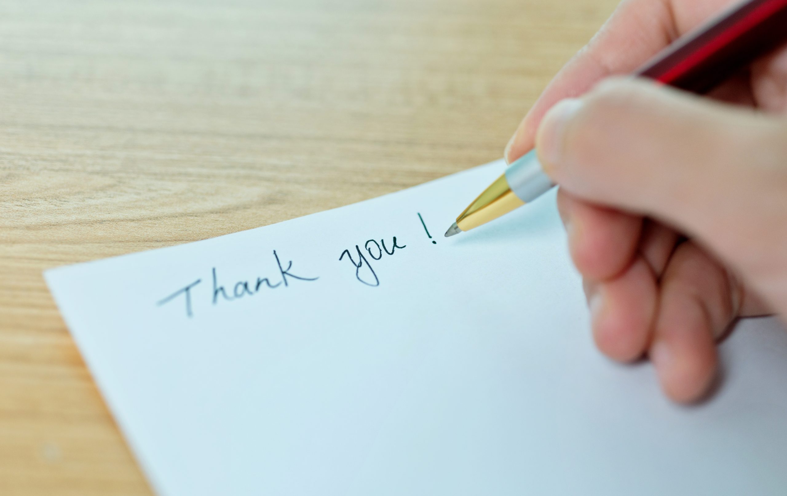 A hand holds a pen that appears to have written the words “Thank you!” on a note card sitting on wood surface. Adobe Stock Images