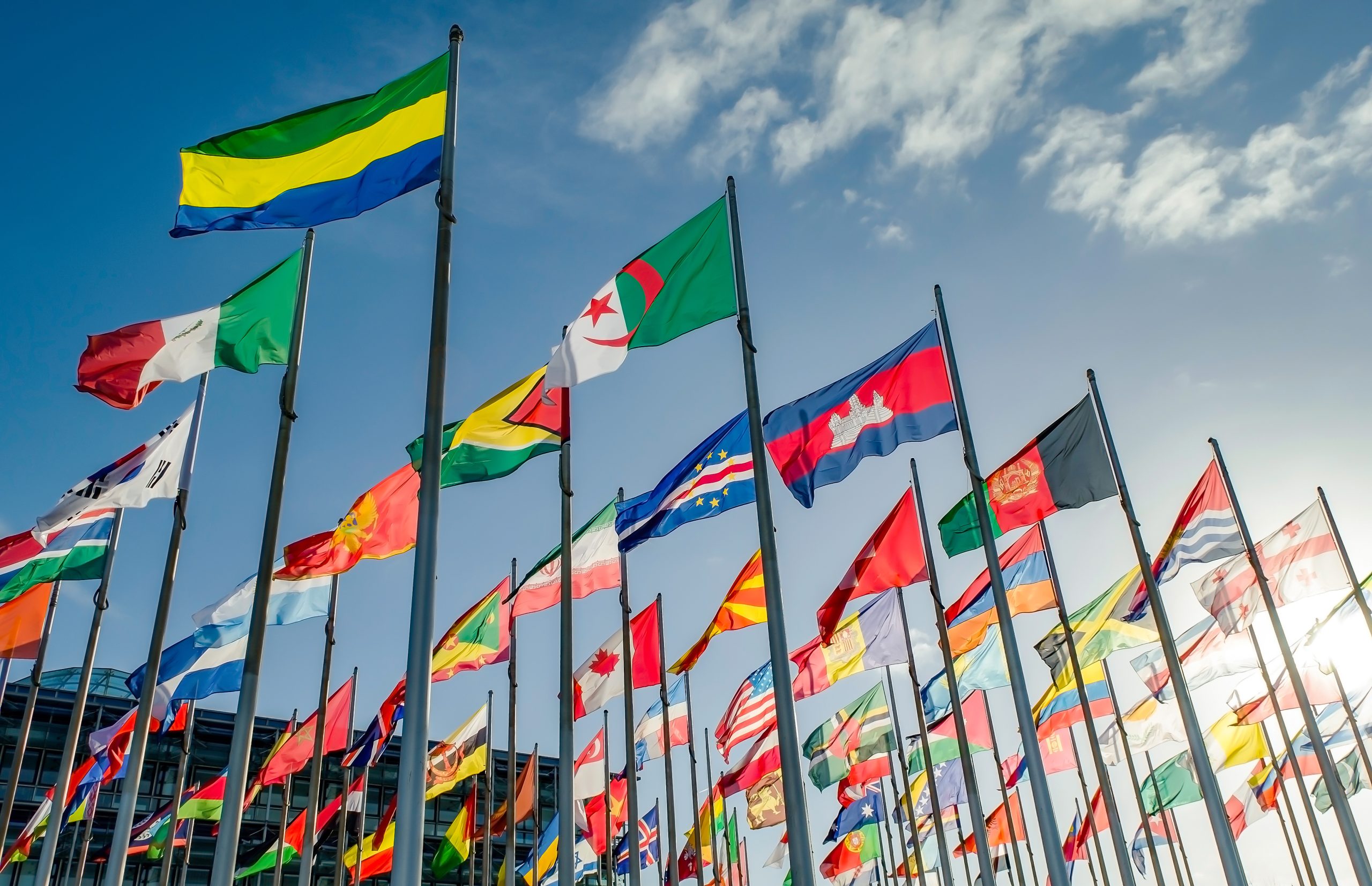 Thirty to forty flags from different countries against a blue sky with puffy white clouds. Adobe Stock Image