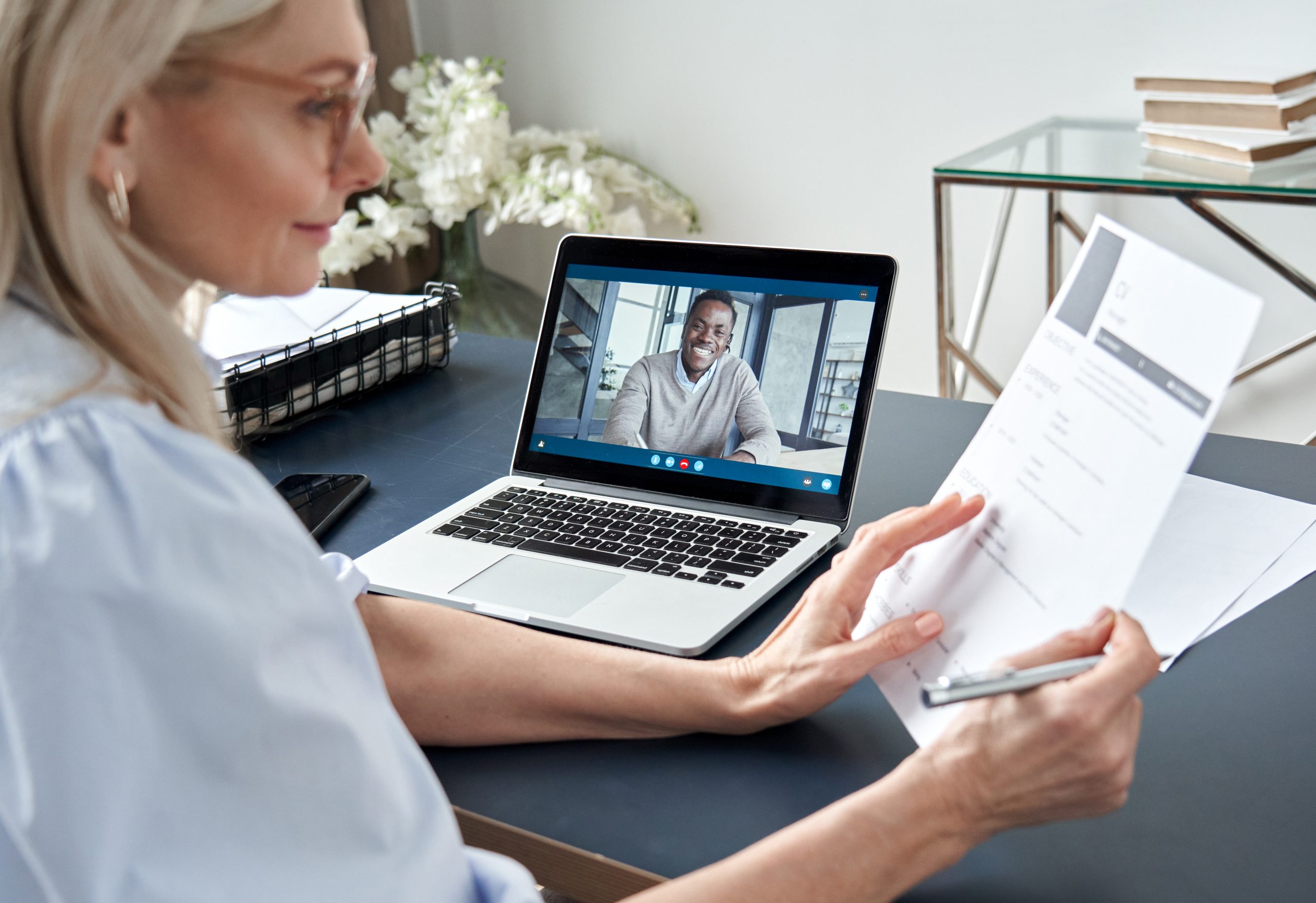 A woman reviews a paper resume sitting in front of a laptop with a man smiling in the video for a video interview. Adobe Stock Image