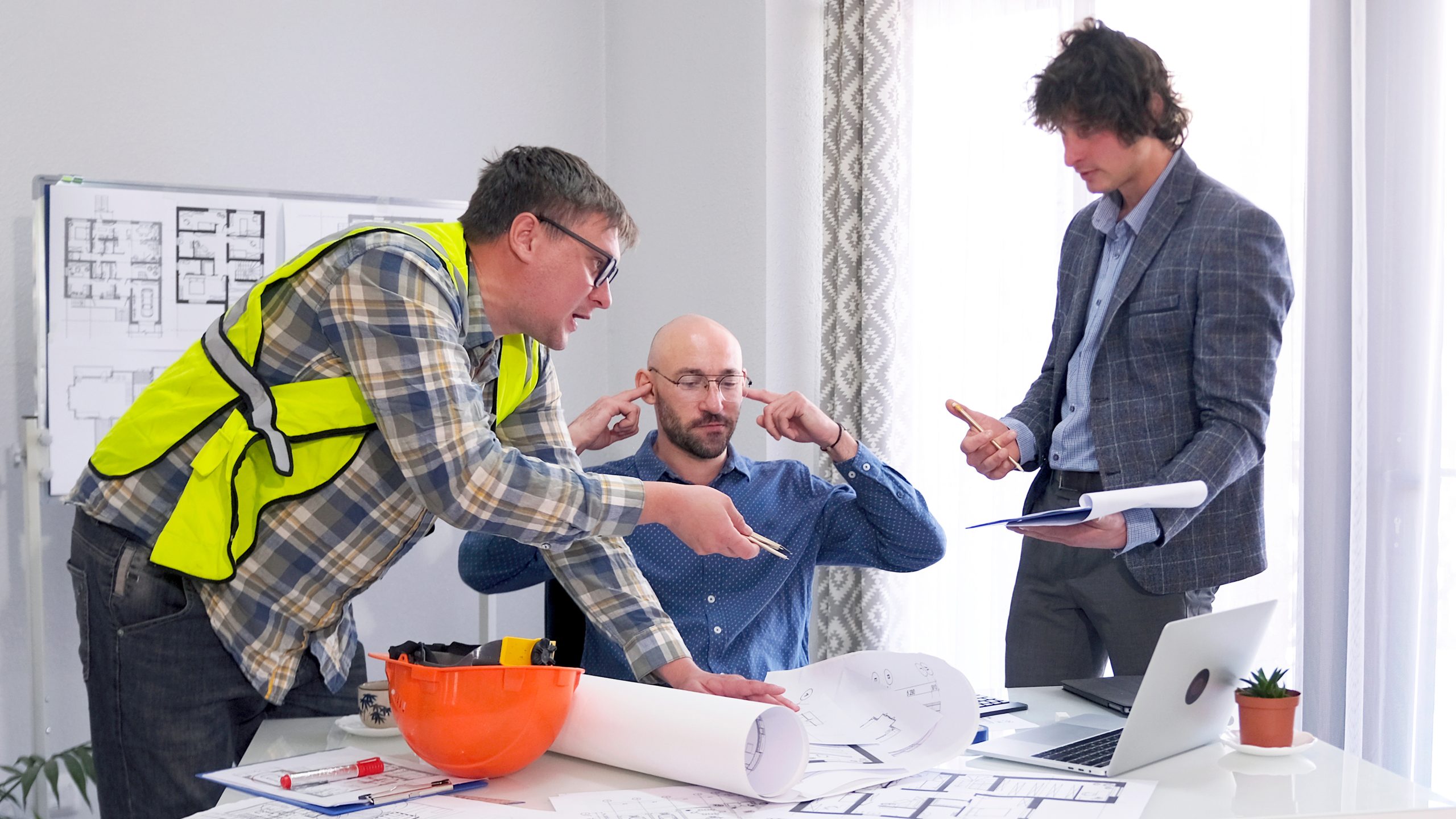 Three people in an office setting engaged in a construction or architectural planning discussion. One person in a high-visibility vest points at architectural plans on a table. Another person sits with fingers in ears, and a third stands holding a notebook and gesturing. A whiteboard with floor plans and a laptop are visible in the background. Adobe Stock Image